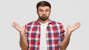 Photo of hesitant unshaven male clasps hands with hesitation, has clueless expression, doubts what to do, dressed in checkered shirt, stands against white background. People and confusement concept